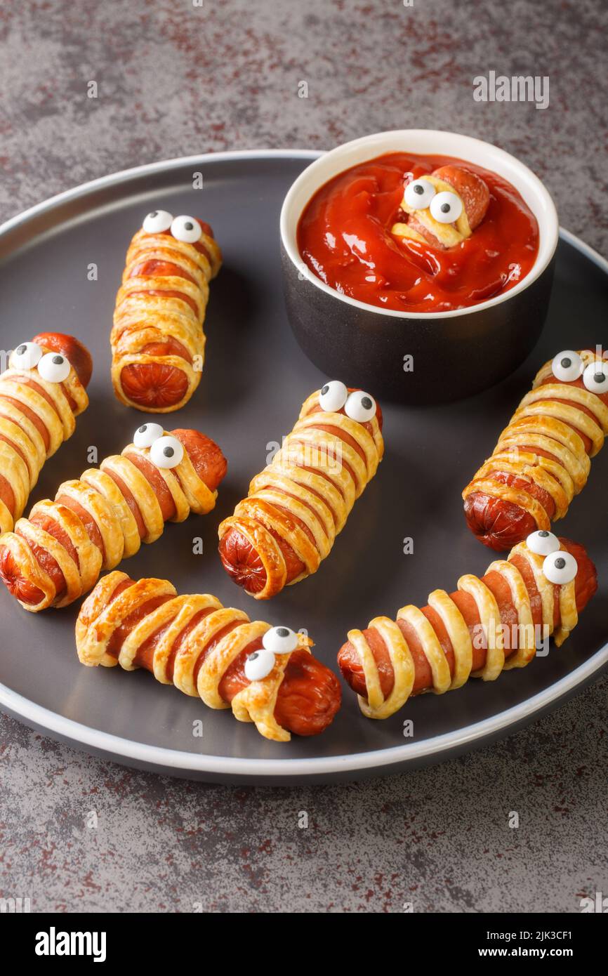 Mummy sausage in dough scary Halloween holiday food with funny eyes and sauce on a plate on the table. vertical Stock Photo