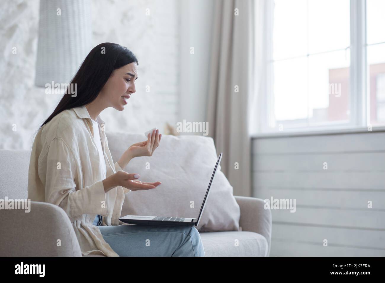 Young beautiful woman alone at home in depression crying, brunette sitting at home and using laptop for online consultation call with psychotherapist Stock Photo