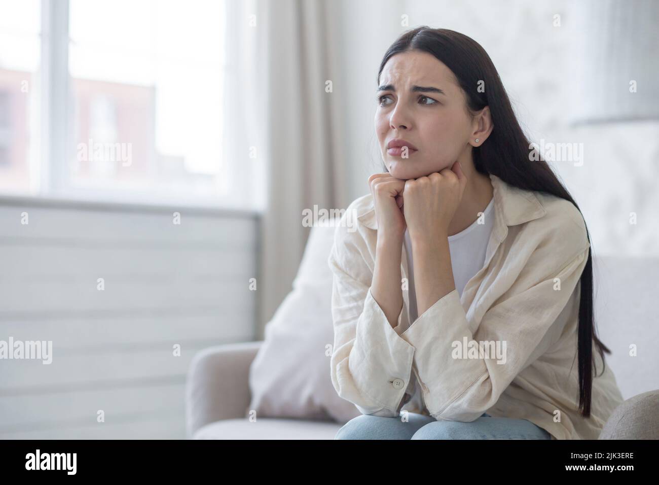 Depressed woman near window sitting on sofa at home, disappointed and sad brunette grieving divorce. Stock Photo