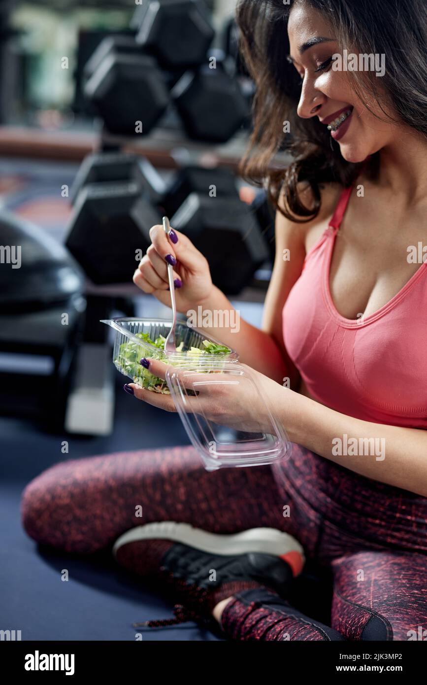Attractive sportswoman in workout clothes eating a healthy salad in a gym. Stock Photo