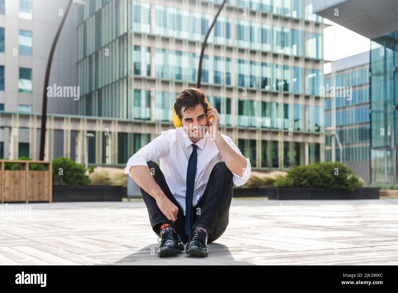 Happy and handsome adult businessman wearing elegant suit sitting on the ground and listening music with headphones to relax Stock Photo