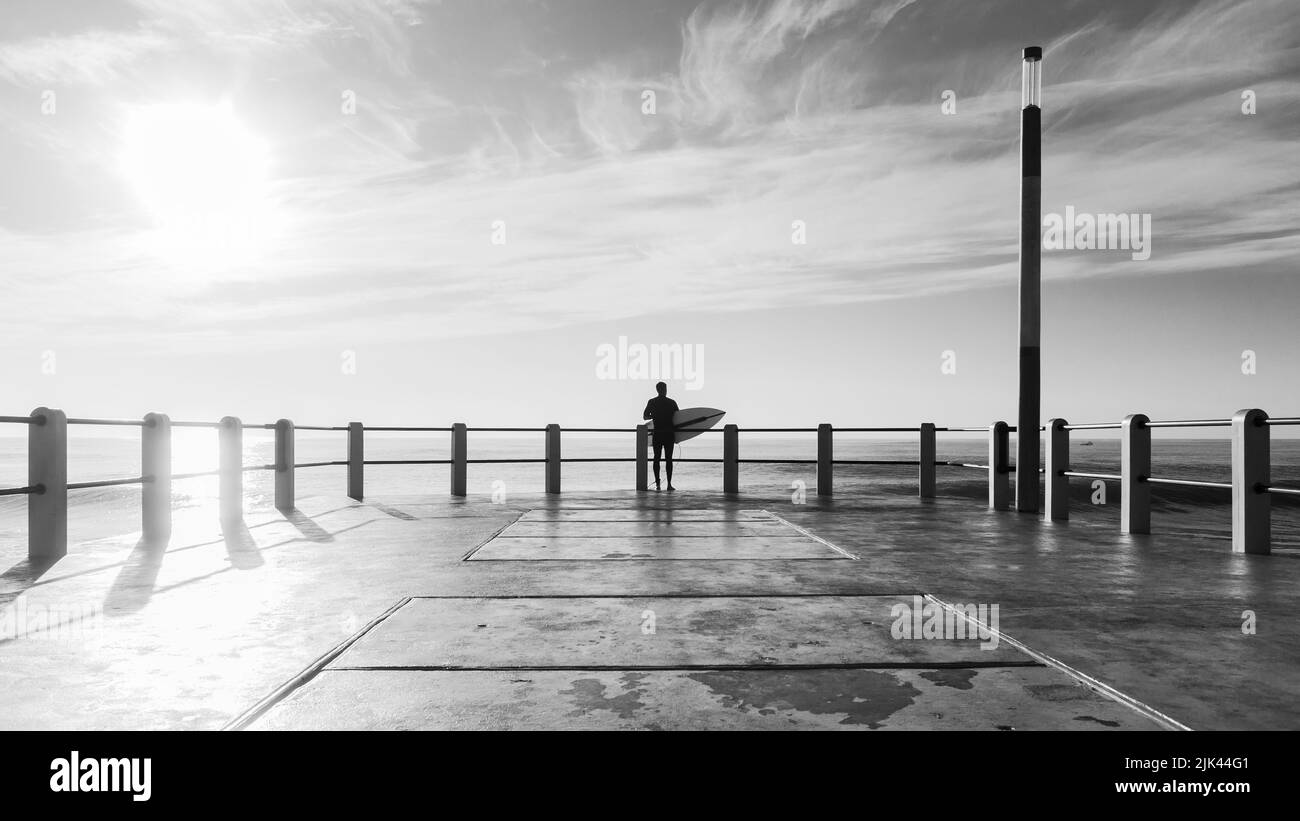Surfer silhouetted standing  end of beach ocean pier jetty about to jump into ocean water going surfing, a rear behind black and white photo. Stock Photo
