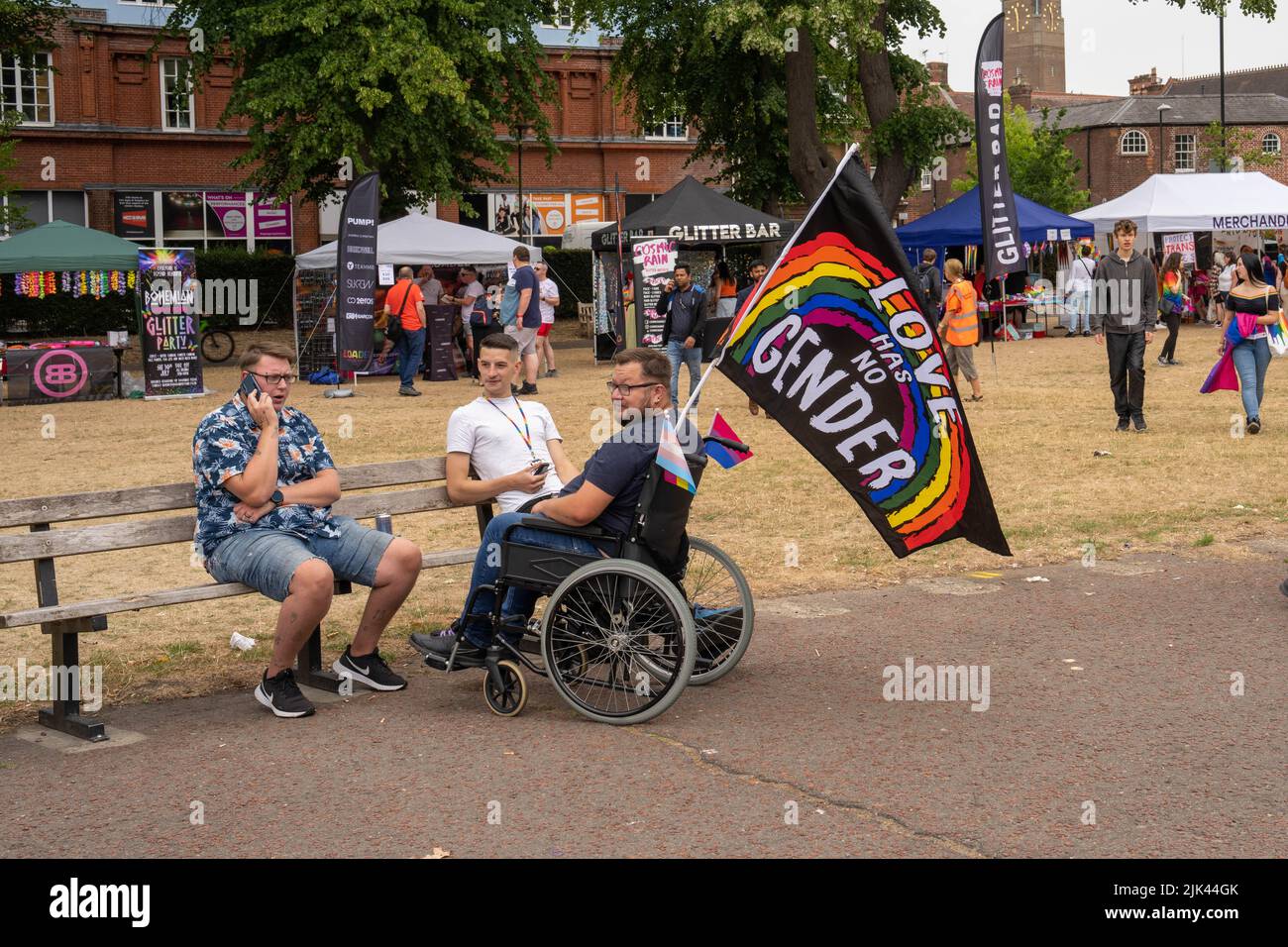 Disabled man with flag stating'Love has no gender'rainbow colours  talking to two men sitting on a bench in Chaplefield gardens Stock Photo