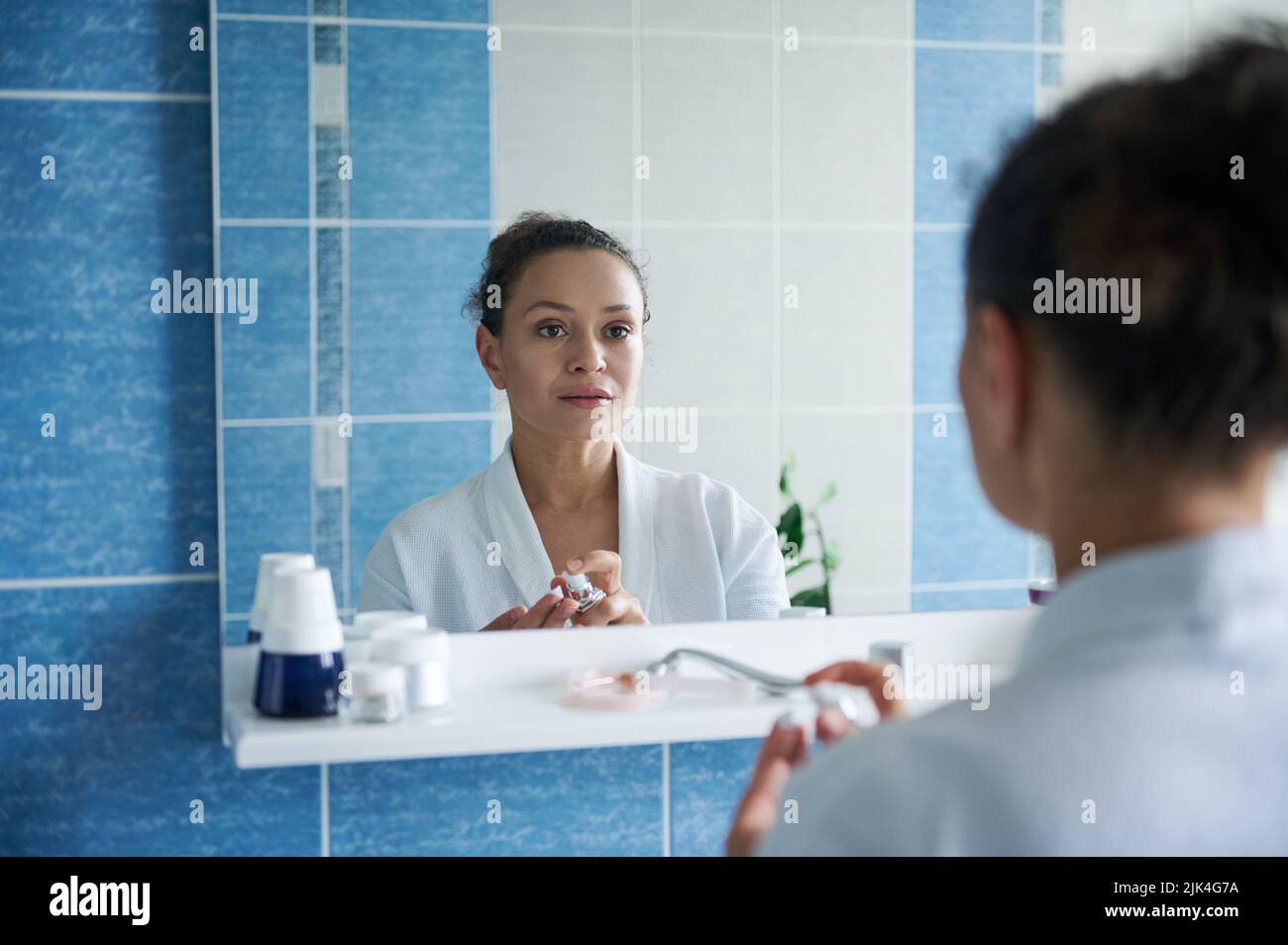 Gorgeous multiethnic woman in white waffle bathrobe looks at her reflection in mirror, applying moisturizer on her hands Stock Photo