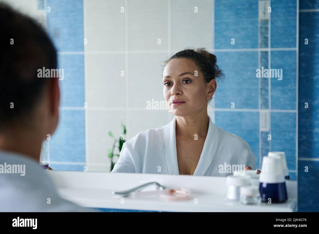 Middle-aged gorgeous woman in bathrobe, looking at her reflection in the bathroom mirror during morning beauty procedure Stock Photo
