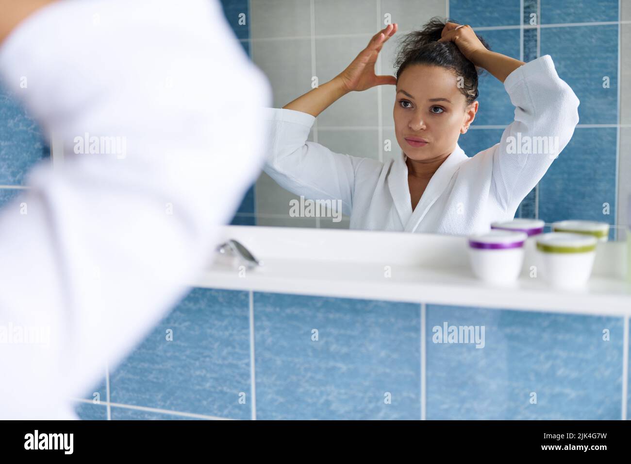 Pretty dark-haired Hispanic woman in white bathrobe ties her ponytail and looks at her reflection in the bathroom mirror Stock Photo