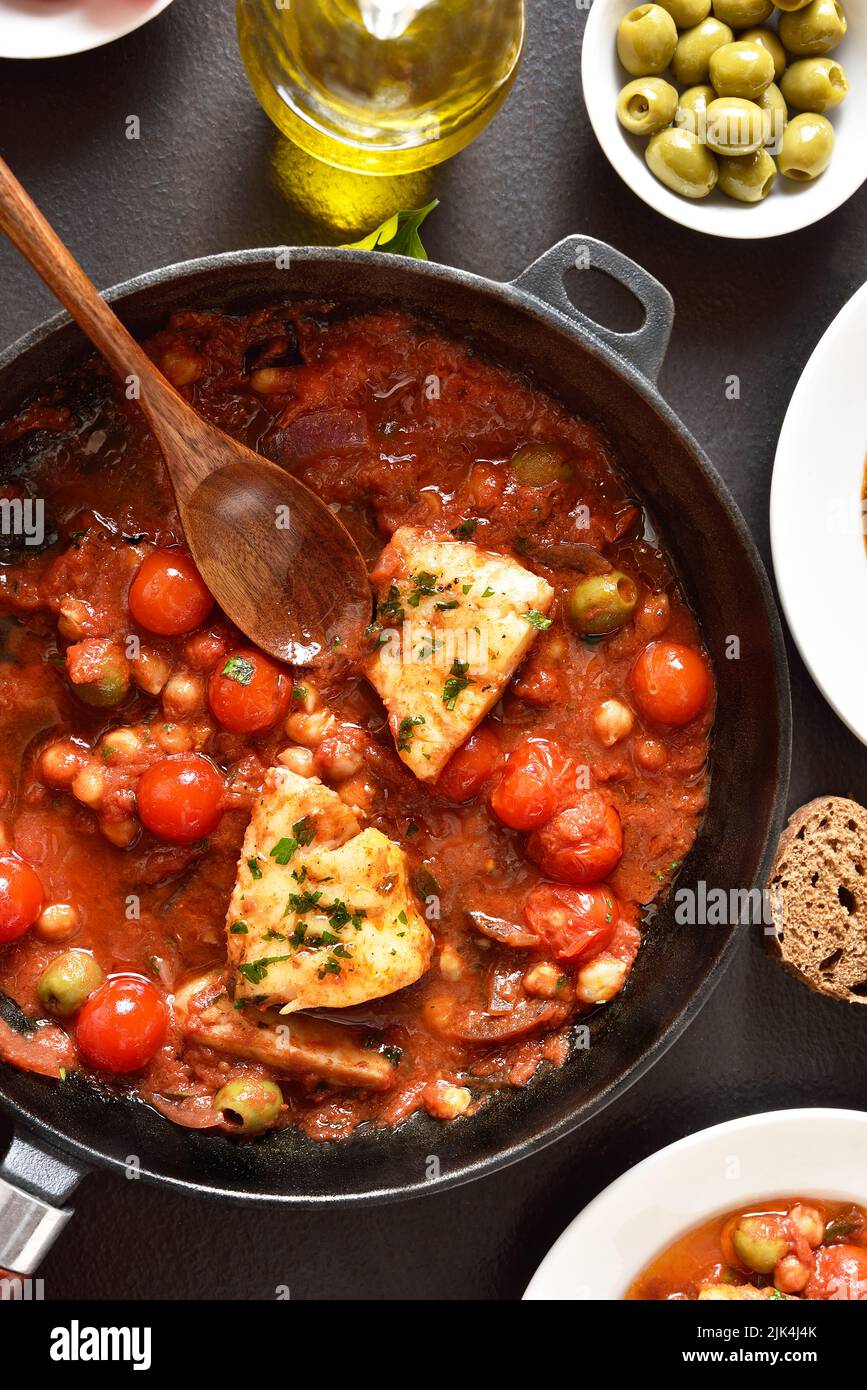 Cod stew with chickpeas, cherry tomatoes and olives in pan over dark stone background. Top view, flat lay Stock Photo