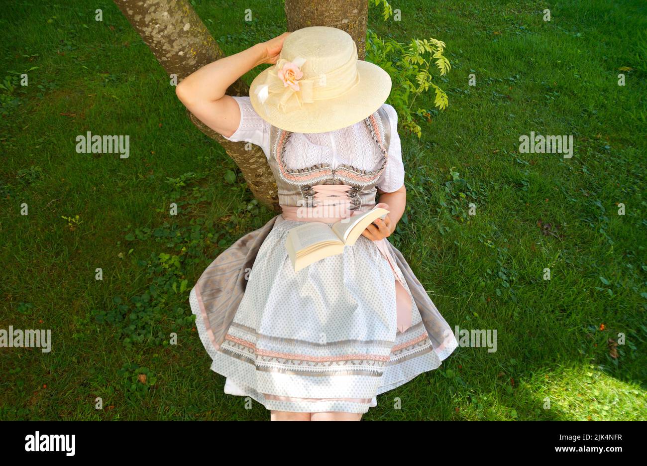 a woman in a beautiful traditional Bavarian dirndl dress (or Tracht) reading a book on the green meadow on a sunny summer day (Munich, Bavaria, German Stock Photo
