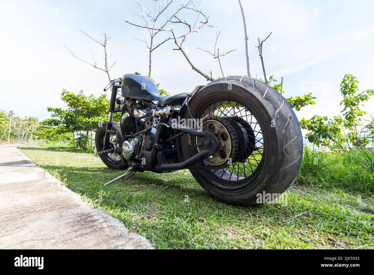 A custom chopper motorbike parked on grass next to a tropical beach. Rat bike, low rider, Honda engine Stock Photo