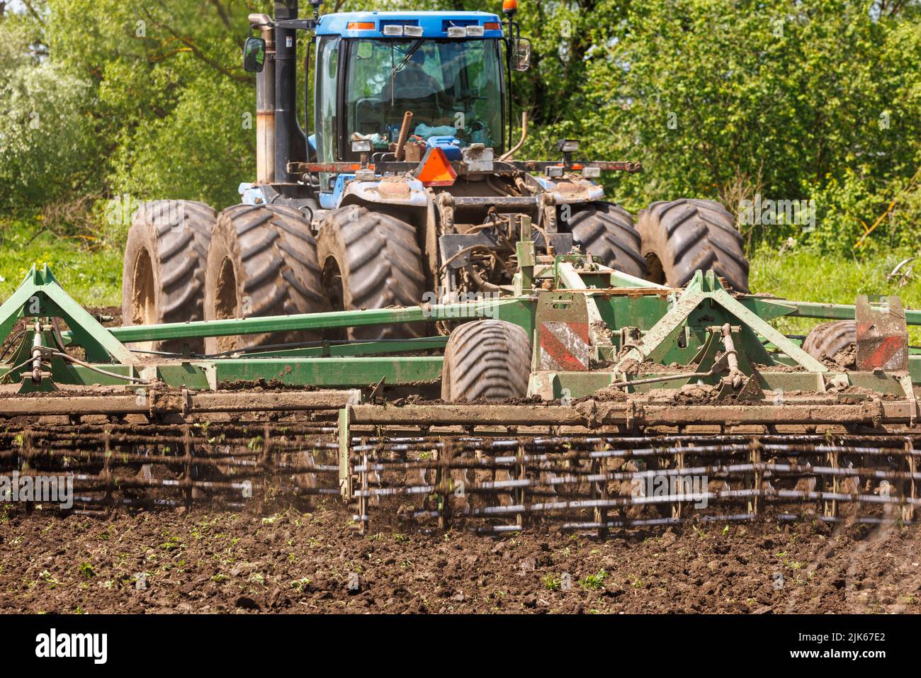 Blue tractor with double wheels pulling disc harrow with roller basket at hot sunny day Stock