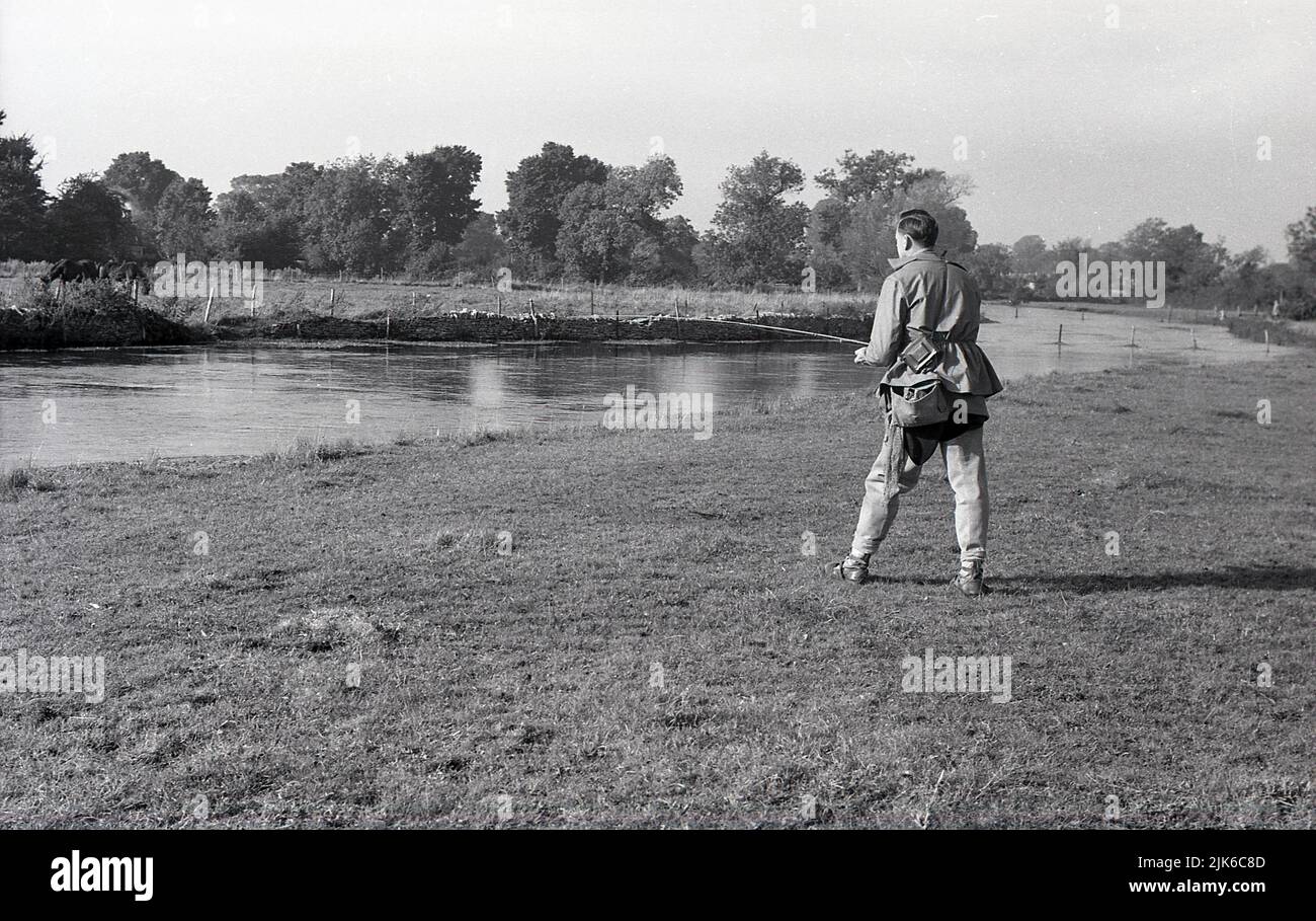 1960s, historical, coarse fishing, outdoors, standing on a wide flat grass riverbank, a man with a rod and wearing the fishing gear of the era, over jacket, waders and shoulder bag, England, UK. Stock Photo