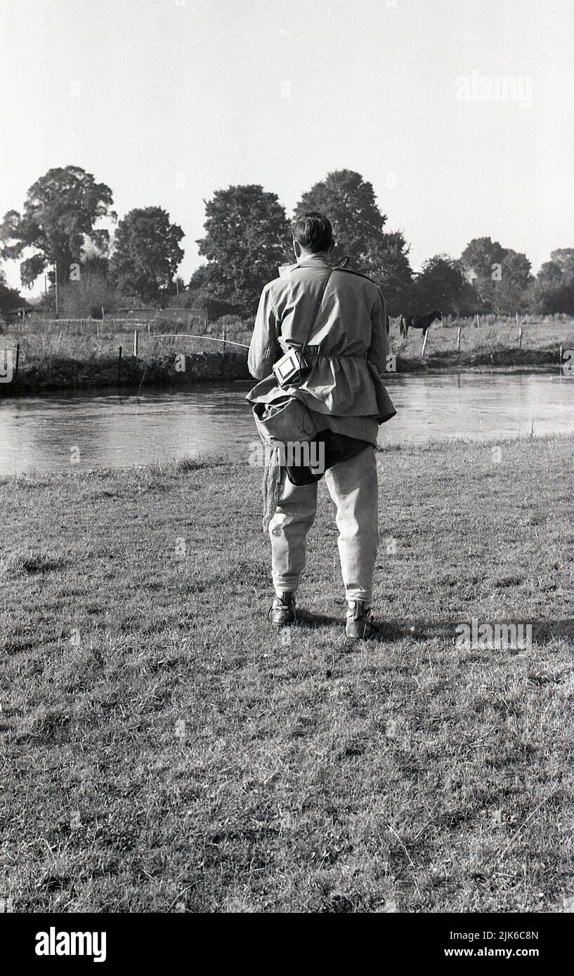 1960s, historical, coarse fishing, outdoors, standing on a wide flat grass riverbank, a man with a rod and wearing the fishing gear of the era, over jacket, waders and shoulder bag, England, UK. Stock Photo