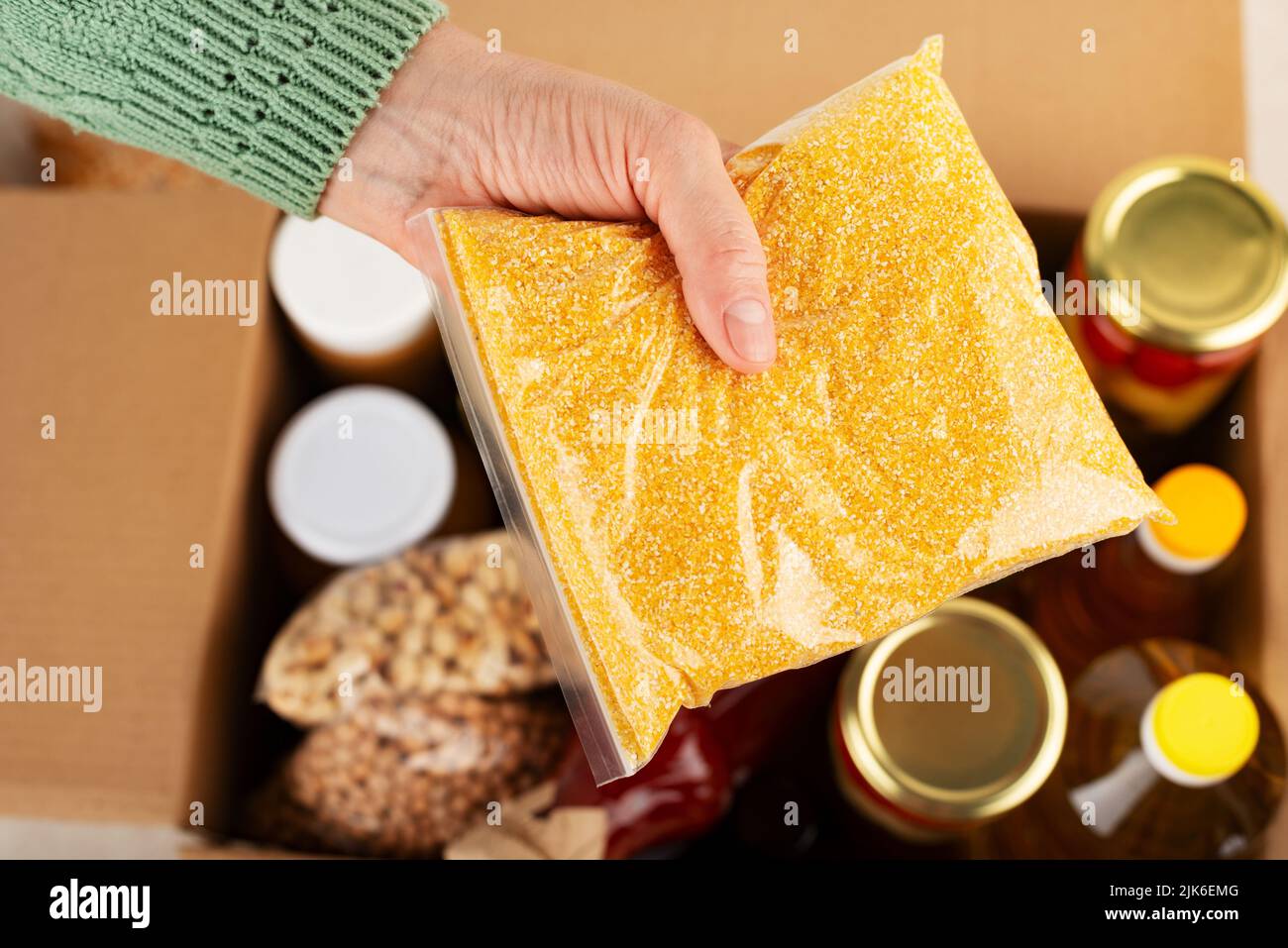 Plastic container with corn grits in female hand on emergency food box background Stock Photo