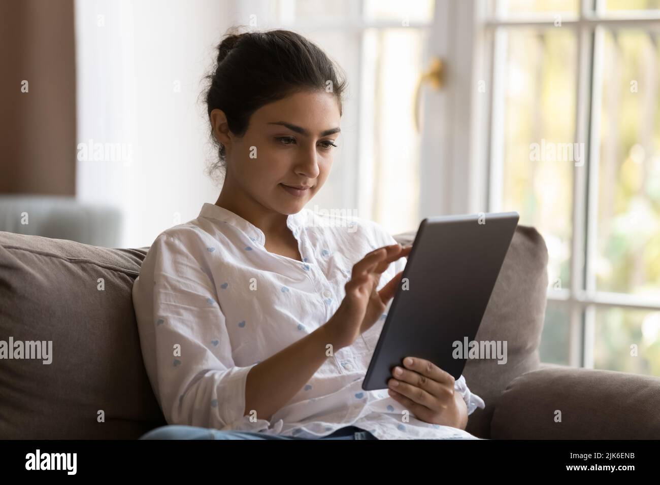 Focused young Indian tablet user woman relaxing on sofa Stock Photo