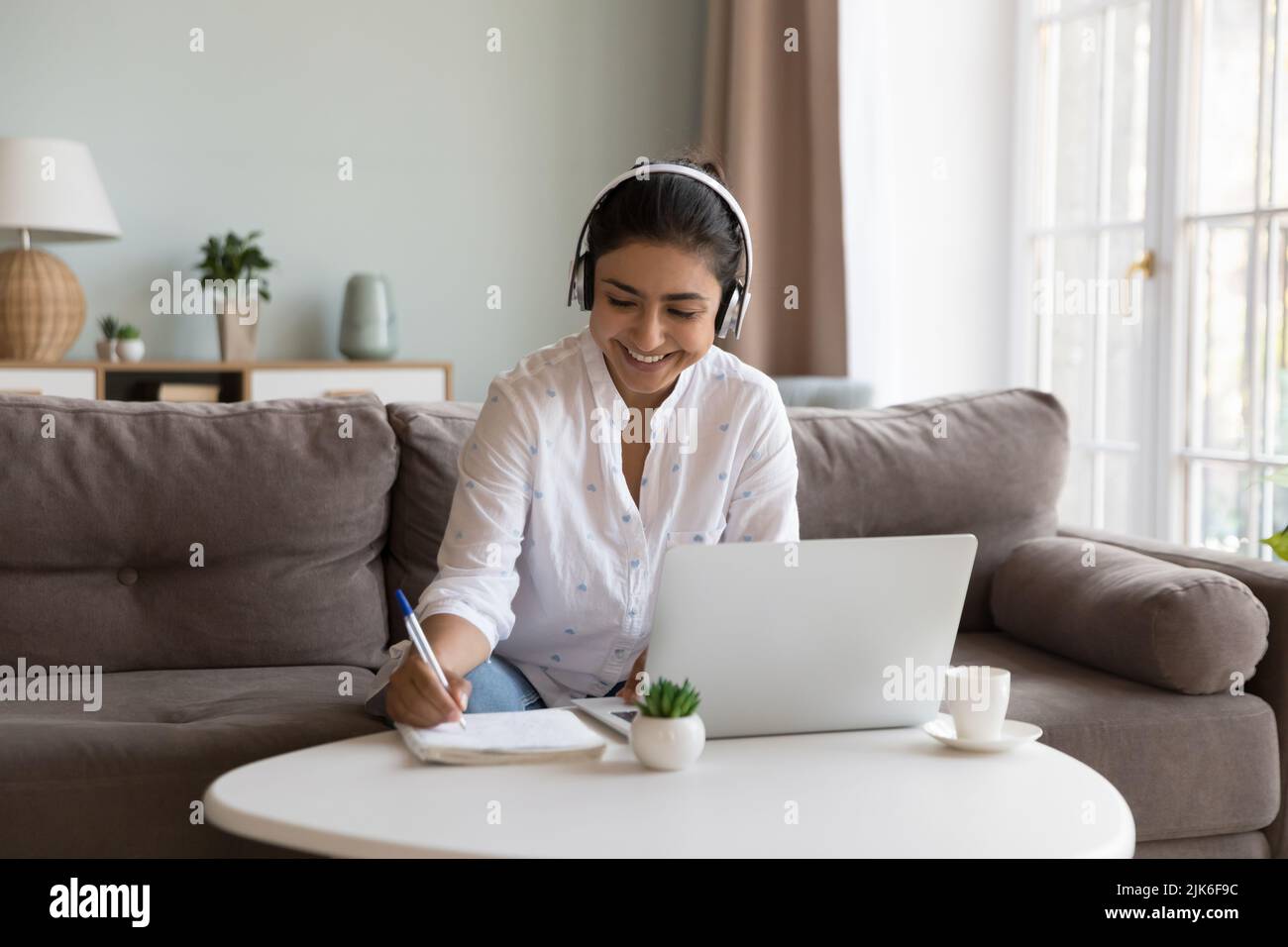 Happy millennial Indian student woman in wireless headphones writing notes Stock Photo