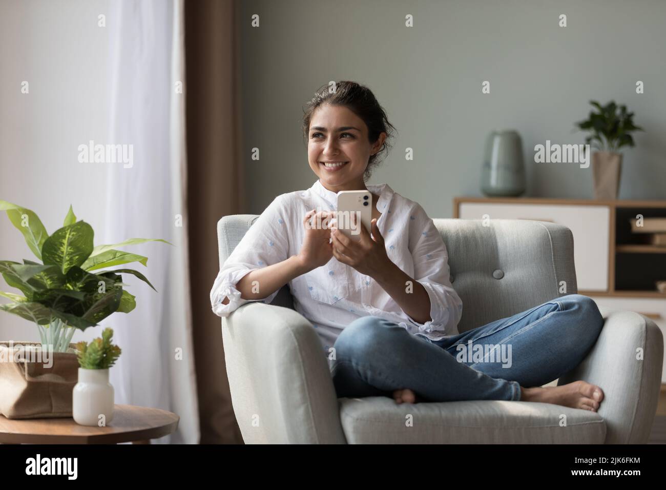 Cheerful beautiful young Indian woman holding cell, sitting in armchair Stock Photo