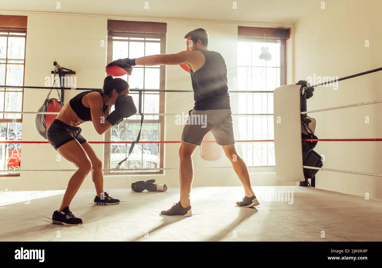 Female boxer ducking a strike from her personal trainer in a boxing ring. Athletic young woman having a boxing training session in a gym. Stock Photo