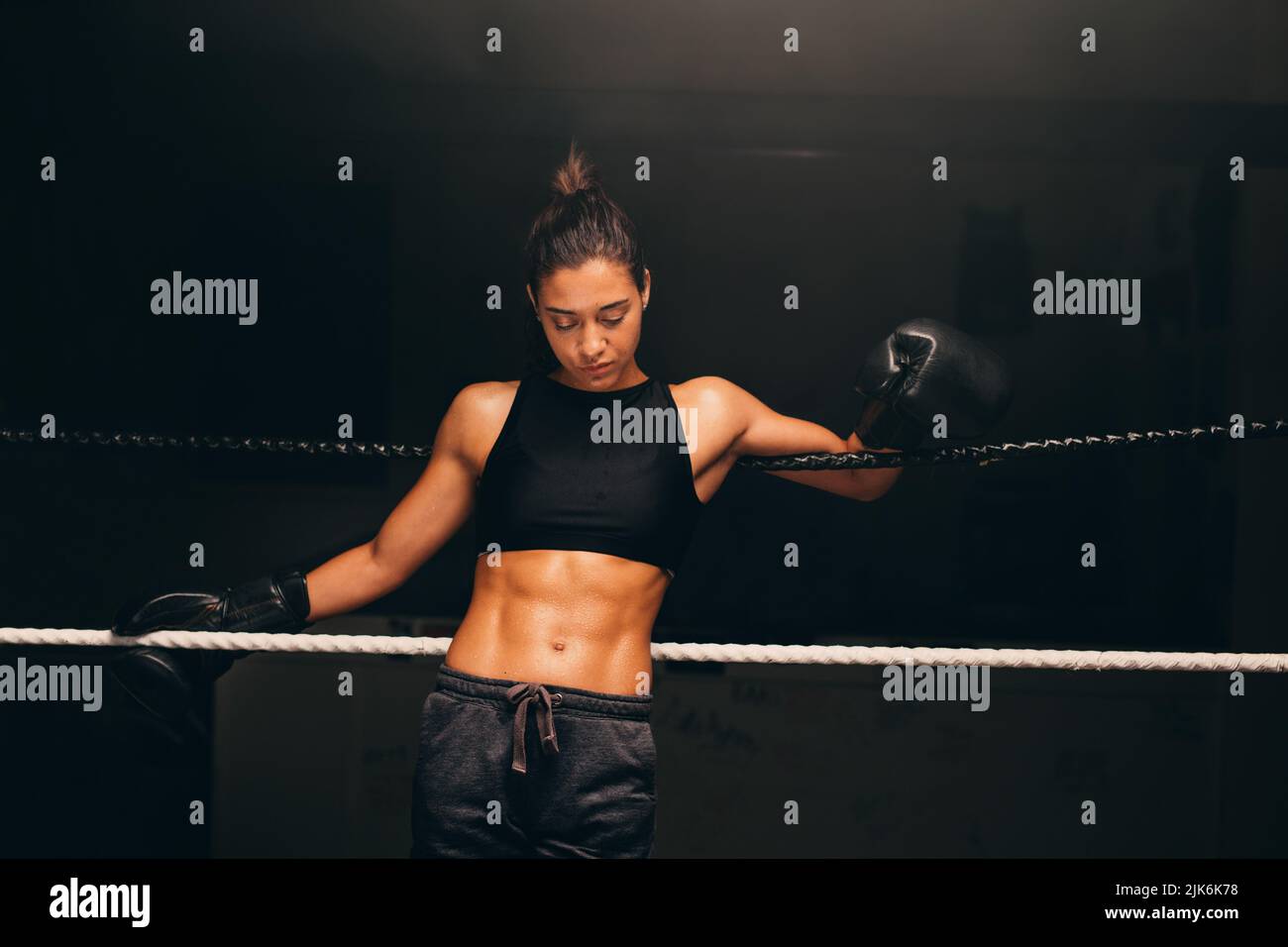 Athletic young woman looking down while standing against the ropes in a boxing ring. Female boxer training in a fitness gym. Stock Photo