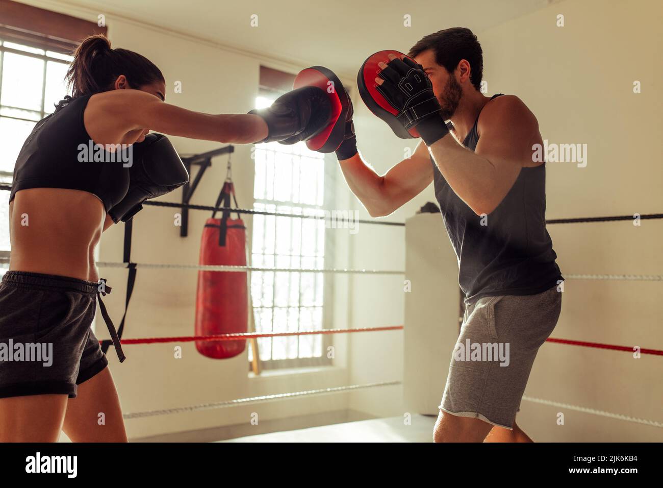 Woman in black shorts throws punch with gloved hands as her instructor holds focus pads in boxing ring Stock Photo