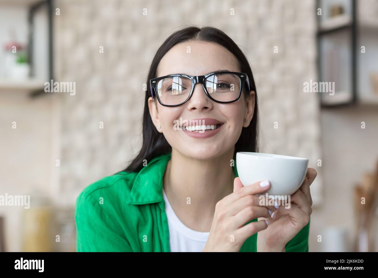 Close-up photo portrait of a young beautiful woman in glasses, holding and drinking a hot drink in the morning, smiling and looking out the window Stock Photo