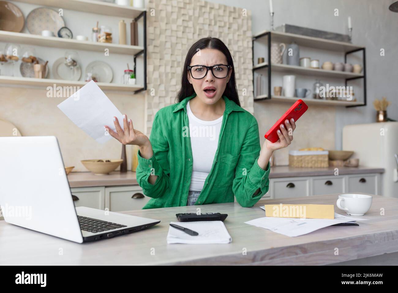 Young beautiful woman in glasses wearing green shirt looking at camera disappointed and angry at homework paper work, holding red phone, brunette calculating household finances in kitchen Stock Photo