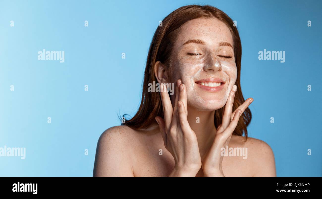 Smiling redhead girl with freckles washing her face, using cleanser