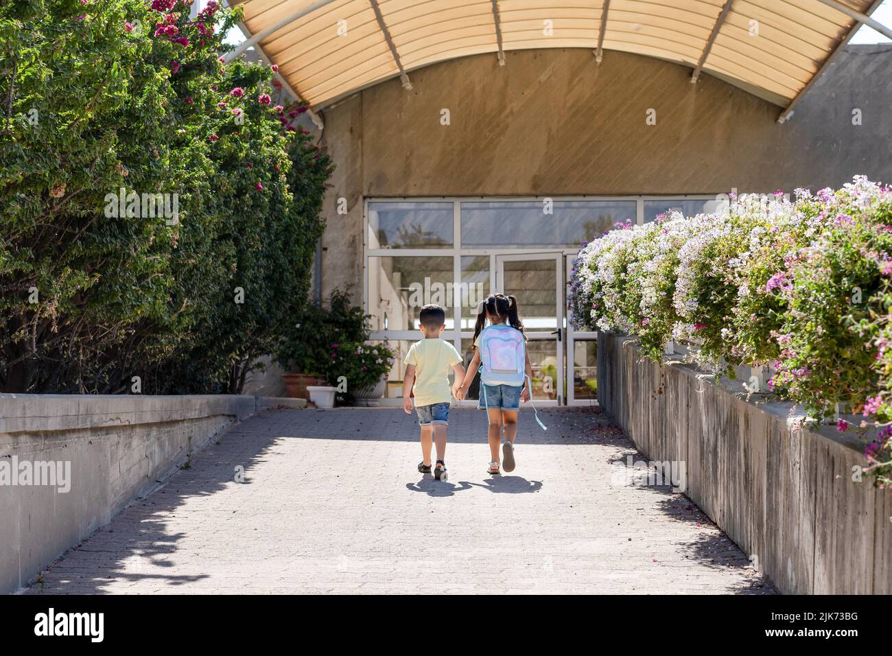 A young children going back to school. Little girl with a tie dye backpack walks to school with her brother. Stock Photo