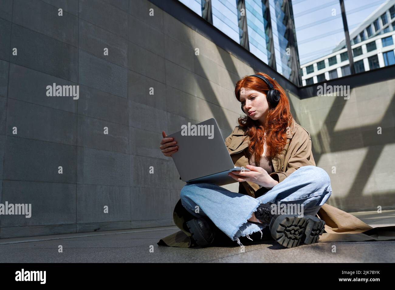 Teen redhead hipster girl wears headphones using laptop in city urban location. Stock Photo