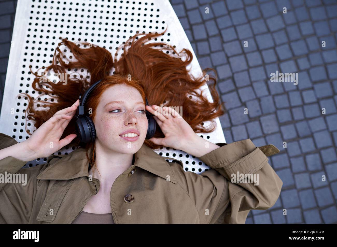 Teen redhead girl wears headphone listening music outdoor looking at copy space. Stock Photo