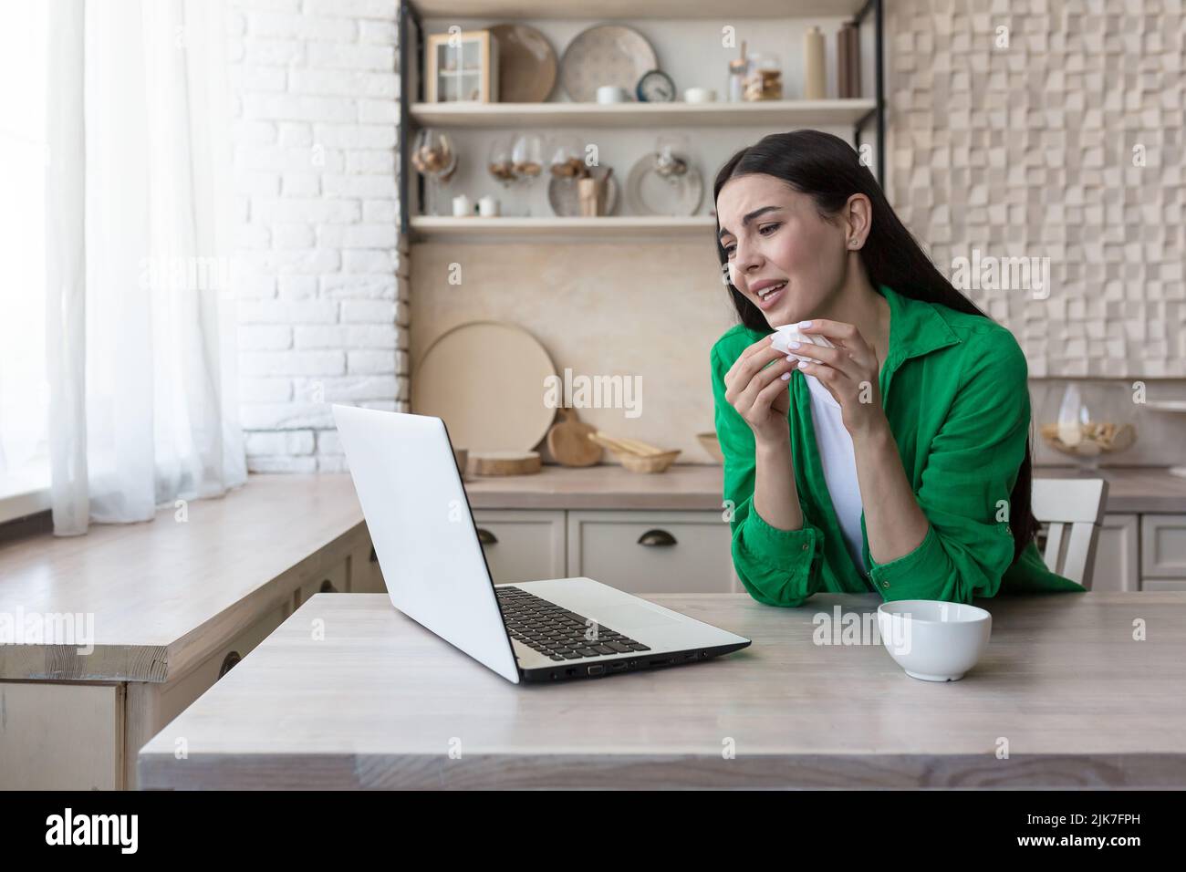 Online consultation with a family psychologist, a young beautiful woman in the kitchen cries in depression, uses a laptop for a video call, consults with a psychotherapist Stock Photo