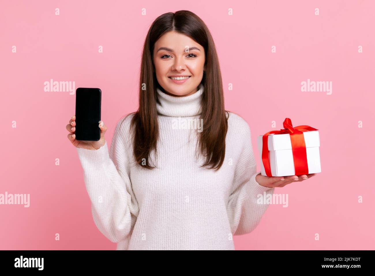 Smiling brunette woman holding wrapped present box and smart phone with blank screen for promotion, wearing white casual style sweater. Indoor studio shot isolated on pink background. Stock Photo
