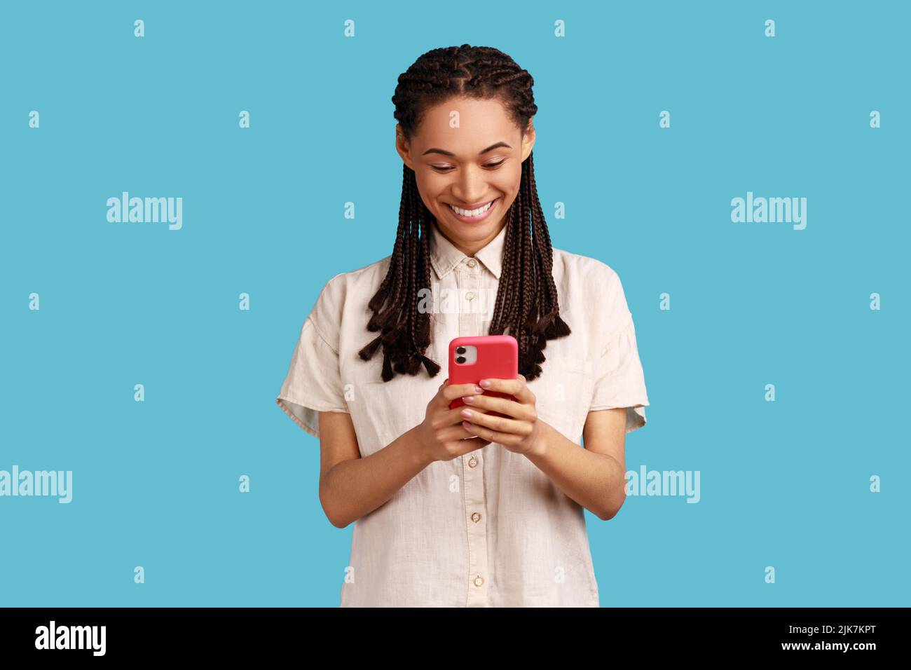 Cheerful woman with black dreadlocks sends text messages via mobile phone, searches gifts for holiday in internet, uses smartphone app, browses webpage. Indoor studio shot isolated on blue background. Stock Photo