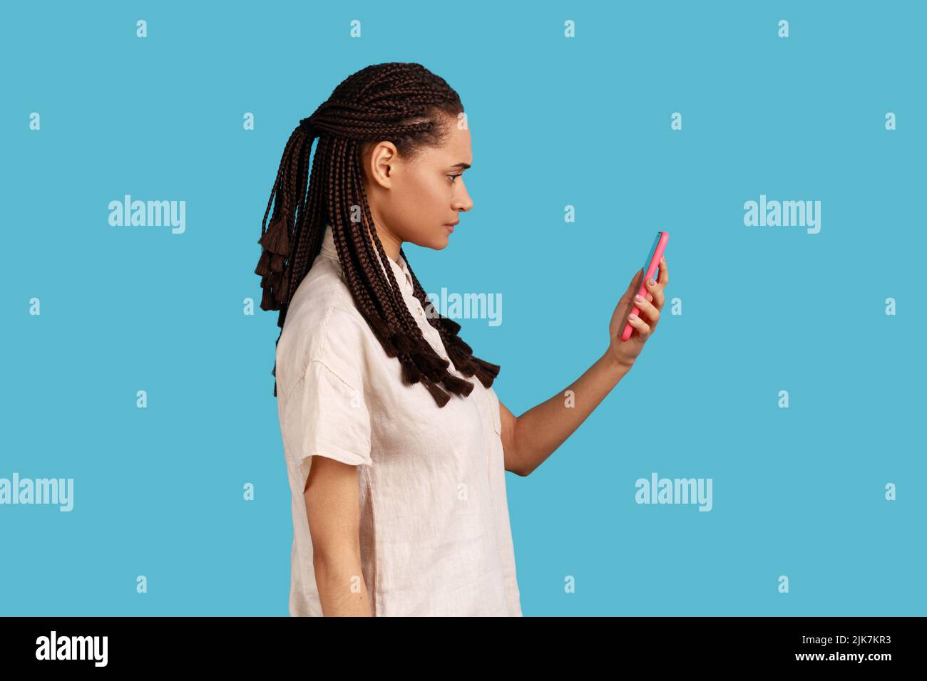 Side view of woman with dreadlocks reads message on modern mobile phone, surfes social media, has concentrated look in display, updates information. Indoor studio shot isolated on blue background. Stock Photo