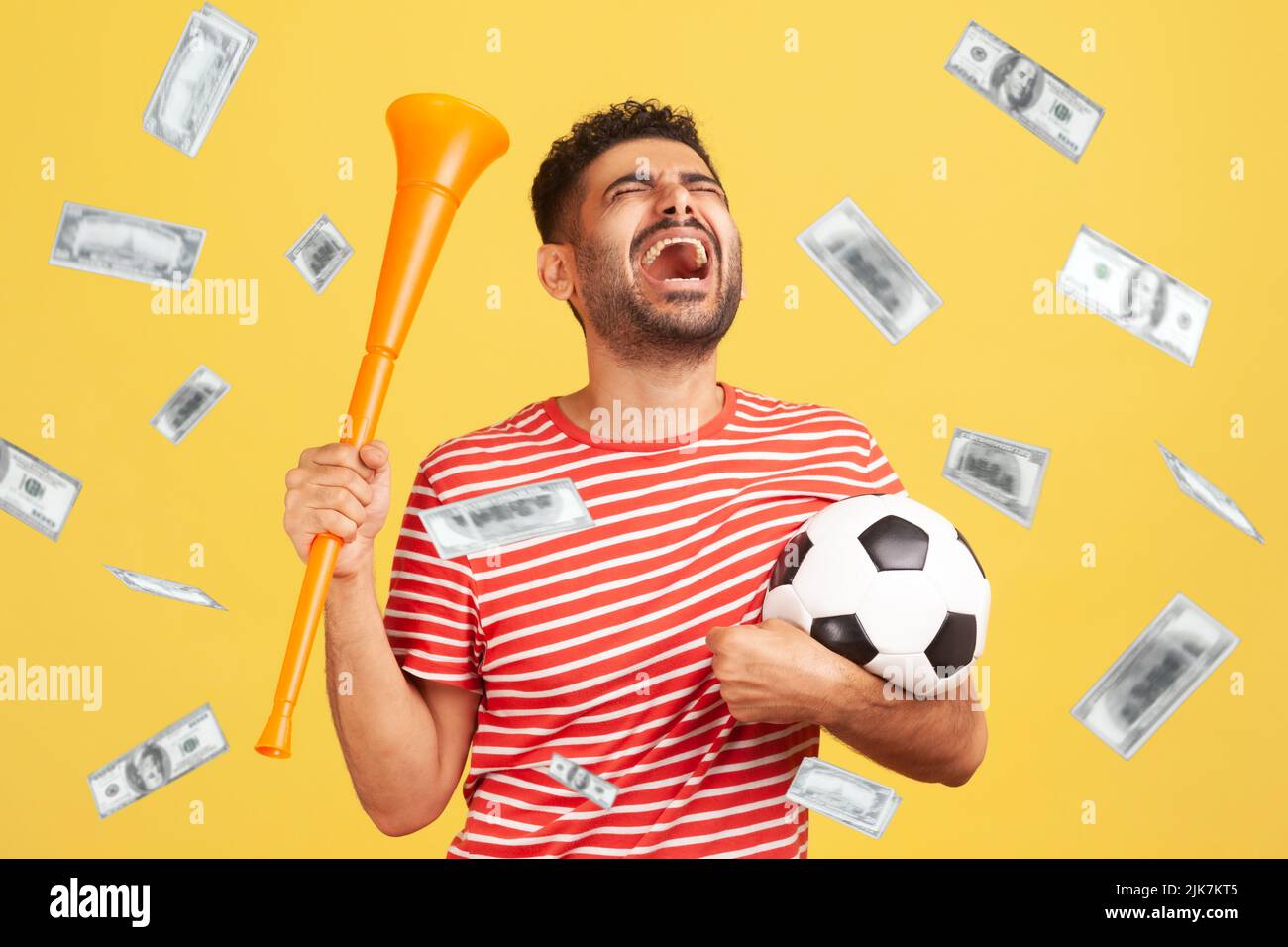 Money rain and rich lucky man extremely happy, holding soccer ball, celebrating victory of favourite football soccer team and his betting. Indoor studio shot isolated on yellow background Stock Photo
