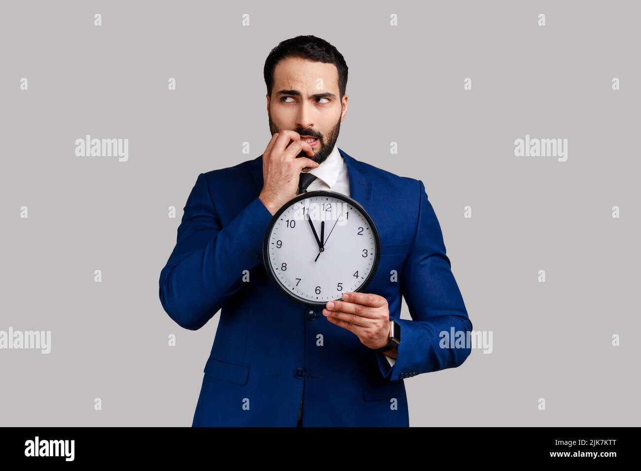 Portrait of attractive nervous bearded man biting nails holding big wall clock, deadline, need hurry up, wearing official style suit. Indoor studio shot isolated on gray background. Stock Photo