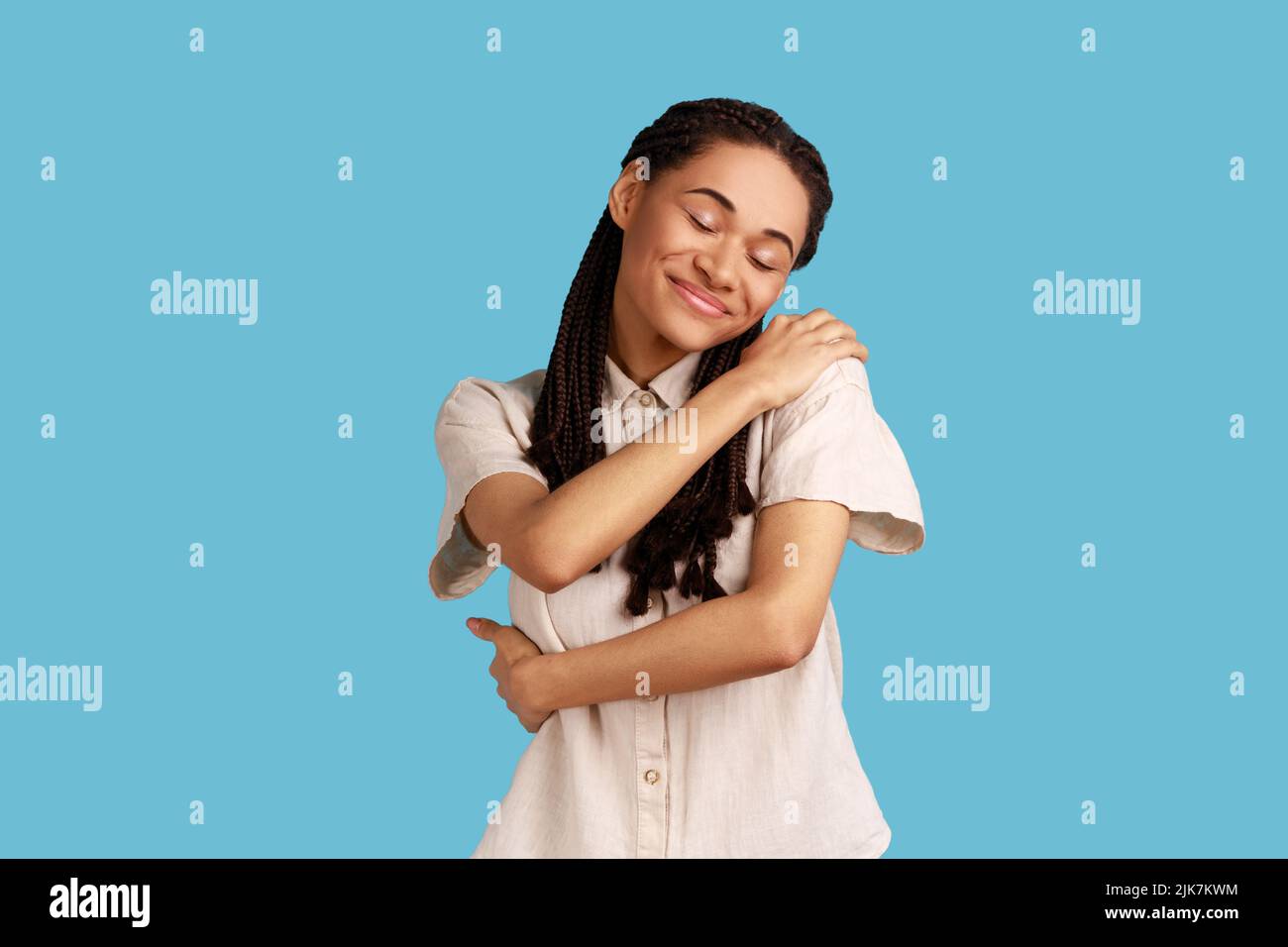 Young cheerful woman with black dreadlocks smiles tenderly, touches shoulders, embraces herself, keeps eyes closed, wearing white shirt. Indoor studio shot isolated on blue background. Stock Photo
