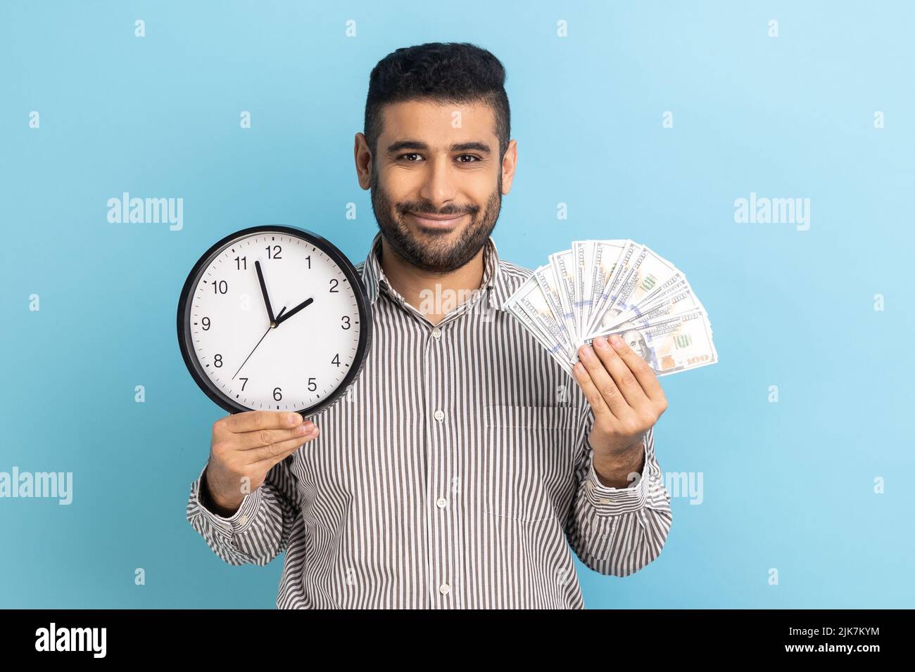 Positive handsome bearded businessman holding big fan of dollar banknotes and wall clock, time is money, expressing happiness, wearing striped shirt. Indoor studio shot isolated on blue background. Stock Photo