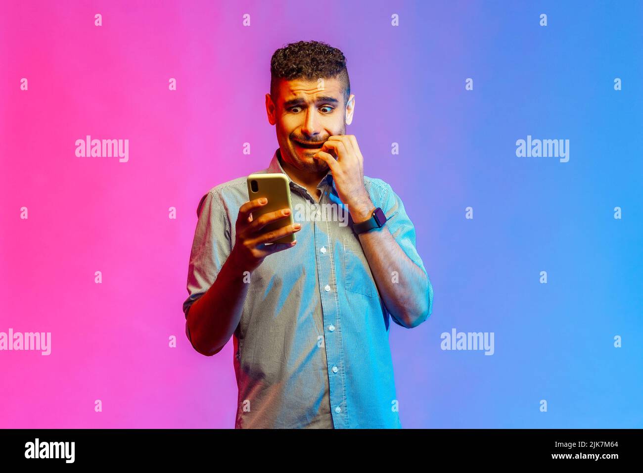 Scared man in shirt standing holding mobile phone, looking at display with fear, biting his finger nails, sees something terrible. Indoor studio shot isolated on colorful neon light background. Stock Photo