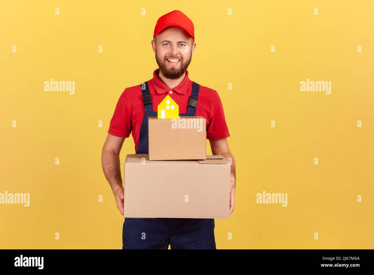 Portrait of courier man in blue coveralls and red T-shirt holding cardboard boxes with paper house on top, movement company, safe delivery. Indoor studio shot isolated on yellow background. Stock Photo