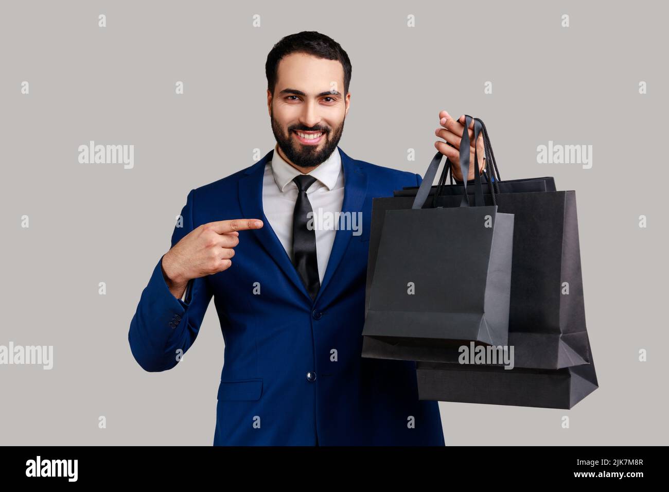 Delighted bearded man pointing finger at paper bags in hand, looking with toothy smile, shopping and sale, wearing official style suit. Indoor studio shot isolated on gray background. Stock Photo