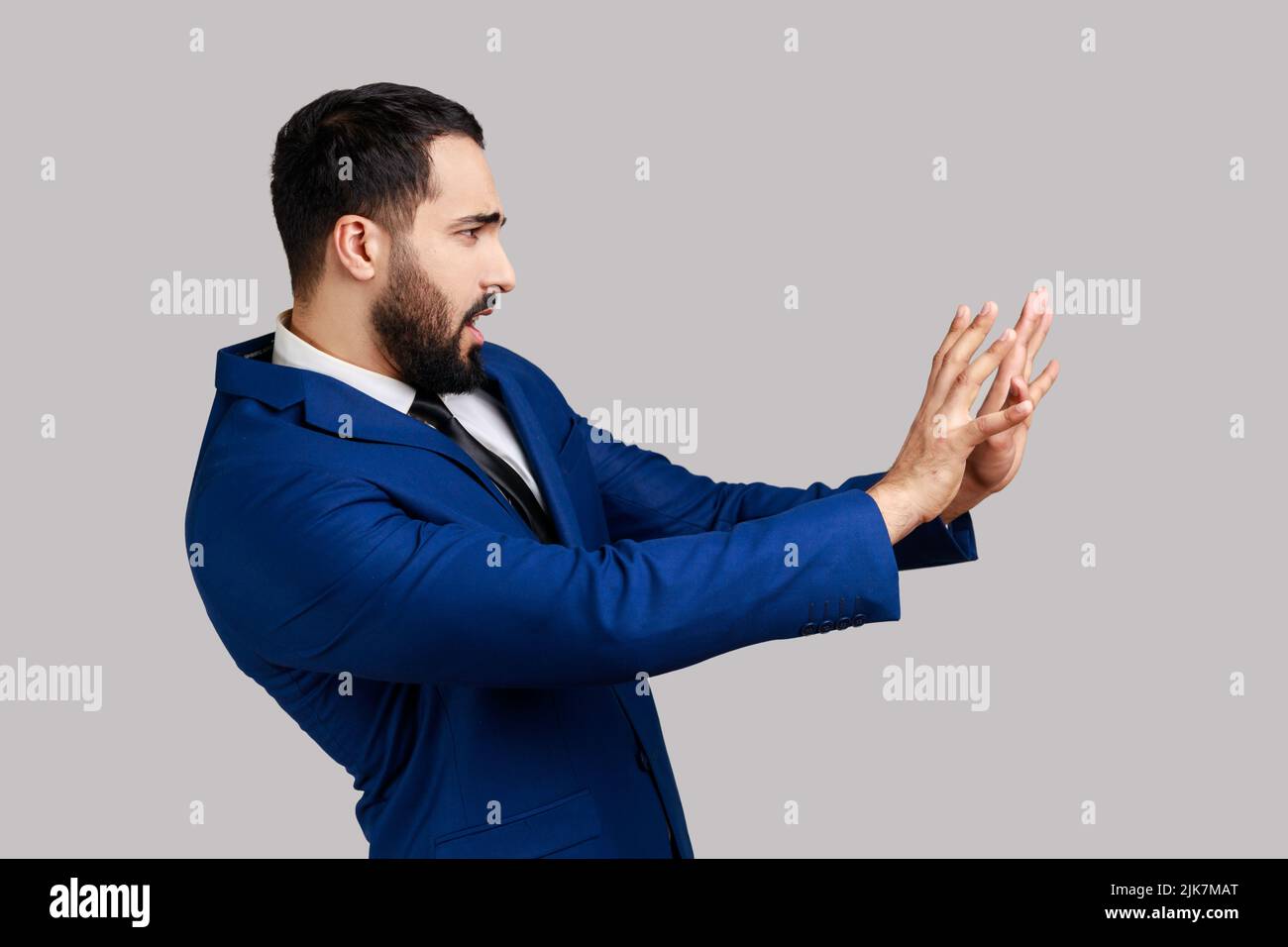 Side view of bearded man raising hands from sudden fear, looking terrified, scared to death, horror facial expression, wearing official style suit. Indoor studio shot isolated on gray background. Stock Photo