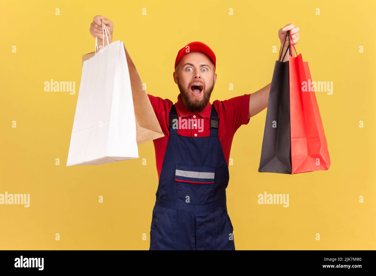 Portrait of amazed bearded courier man in blue uniform raised arms with shopping bags, big sale, bringing order, looking at camera with big eyes. Indoor studio shot isolated on yellow background. Stock Photo