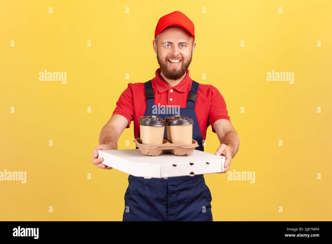 Portrait of smiling professional courier wearing blue overalls standing with pizza box and coffee in disposable cup, fast food delivery service. Indoor studio shot isolated on yellow background. Stock Photo