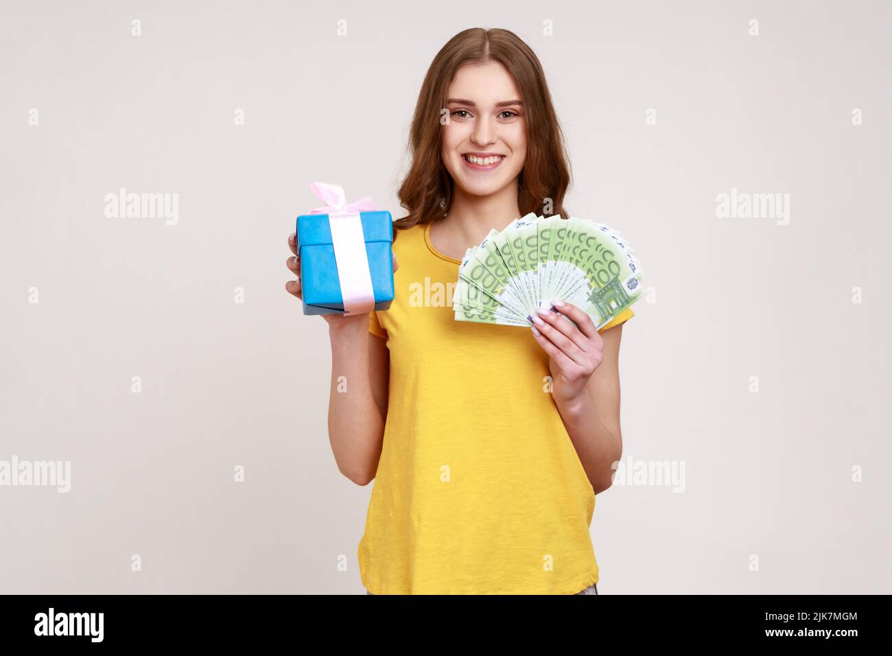 Gift shopping. Happy teenager girl in yellow T-shirt holding present box and euro money banknotes, satisfied with purchase, cashback and bank loan. Indoor studio shot isolated on gray background. Stock Photo