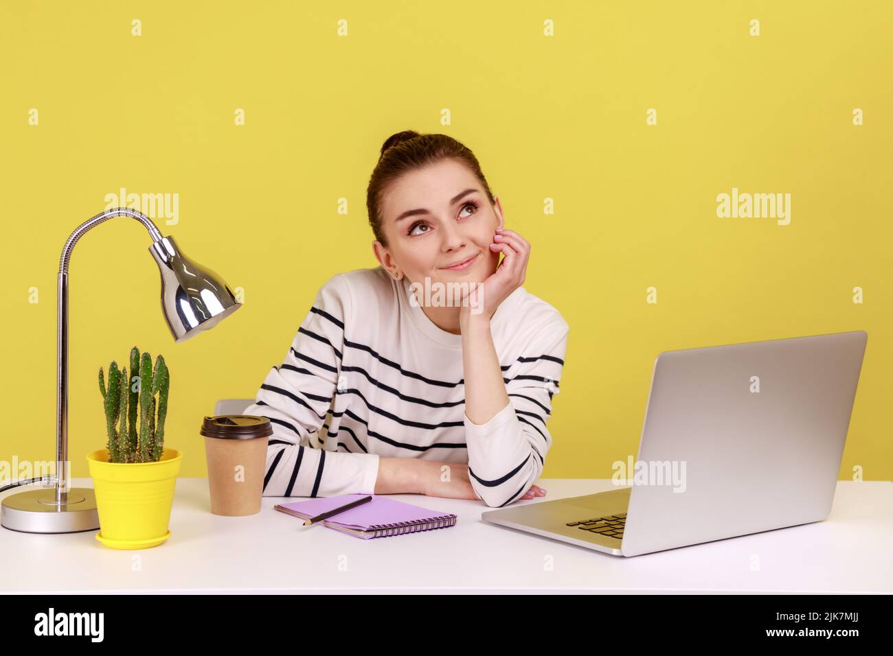 Happy woman dreaming of vacation, resting on break, relaxing with thoughtful meditative expression while working at home office. Indoor studio studio shot isolated on yellow background. Stock Photo