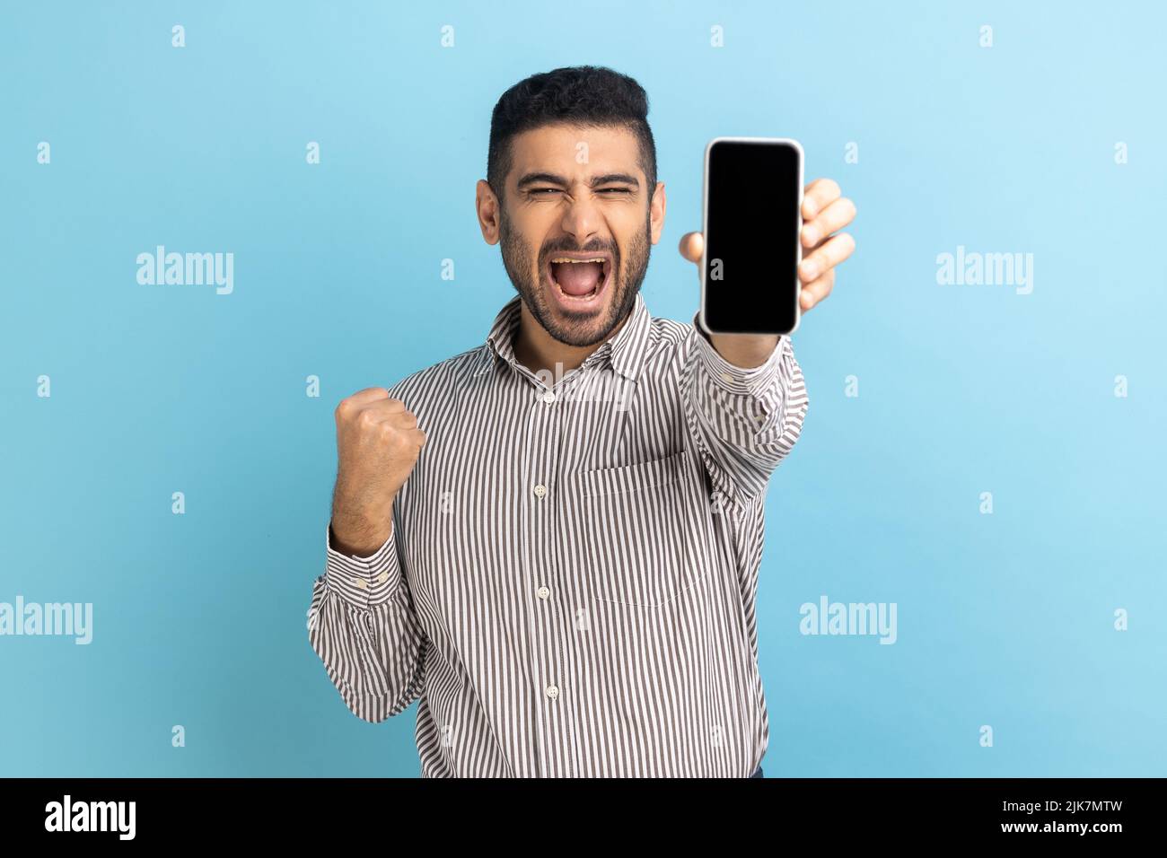 Happy businessman with beard holding smartphone and smiling making yes gesture, celebrating online lottery or giveaway victory, wearing striped shirt. Indoor studio shot isolated on blue background. Stock Photo