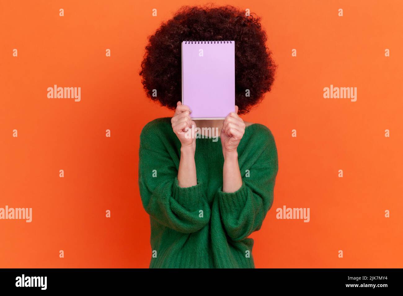 Portrait of unknown woman with Afro hairstyle wearing green casual style sweater standing holding organizer, hiding her face behind paper notebook. Indoor studio shot isolated on orange background. Stock Photo