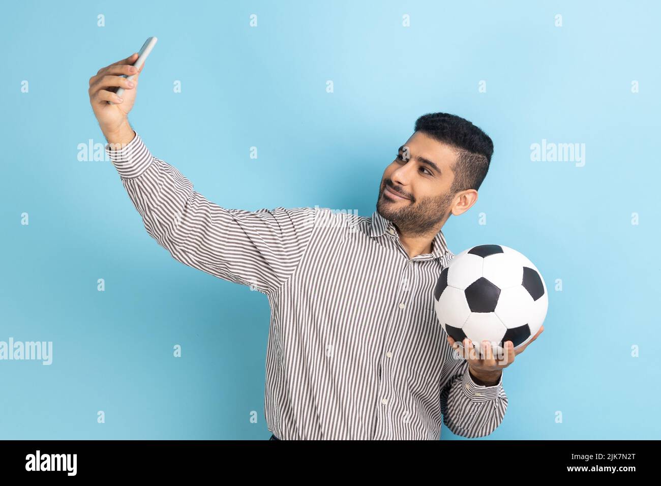 Young adult attractive businessman making selfie, having video call or broadcasting livestream with ball in hand, wearing striped shirt. Indoor studio shot isolated on blue background. Stock Photo