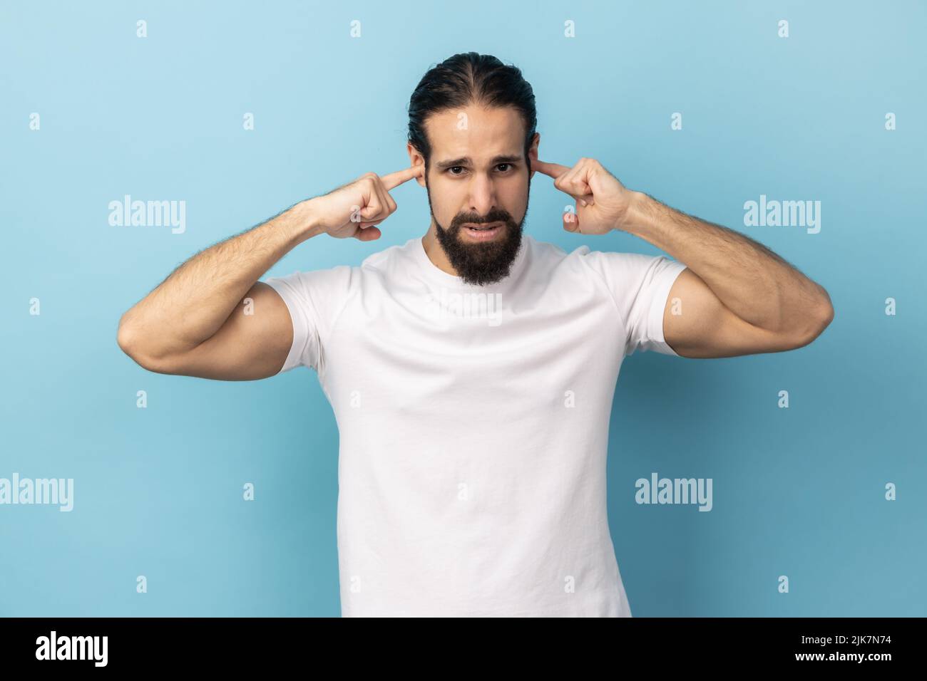 I don't want to here this. Portrait of man with beard wearing white T-shirt standing and holding fingers on his ear, unpleasant sounds. Indoor studio shot isolated on blue background. Stock Photo