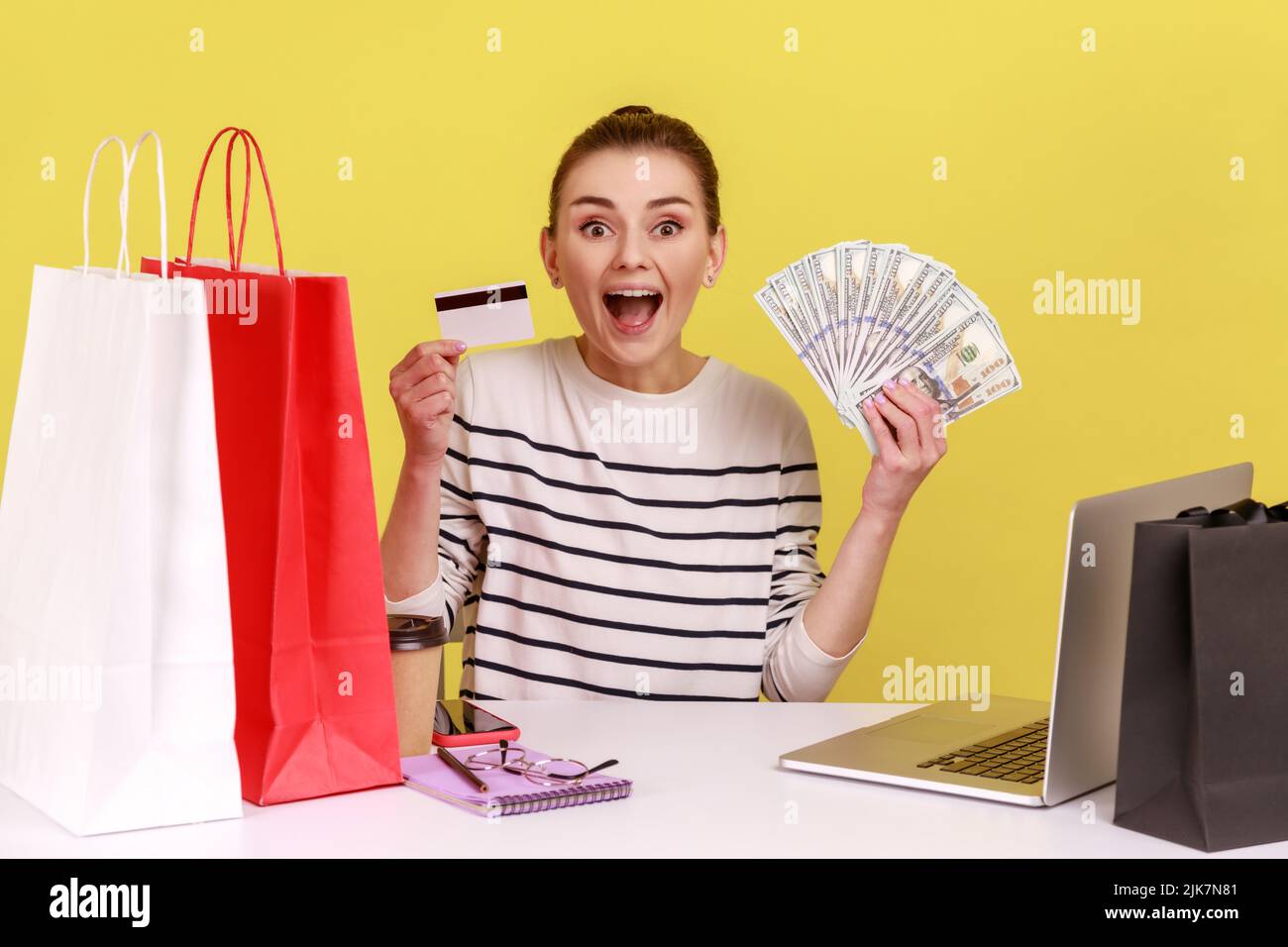 Bank loan, electronic money. Amazed woman sitting office workplace with laptop, holding dollars and credit card, looking at camera with smile. Indoor studio studio shot isolated on yellow background. Stock Photo